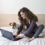 Attractive woman sits on bed with a Jack Russell terrier using a computer to write a pet resume.