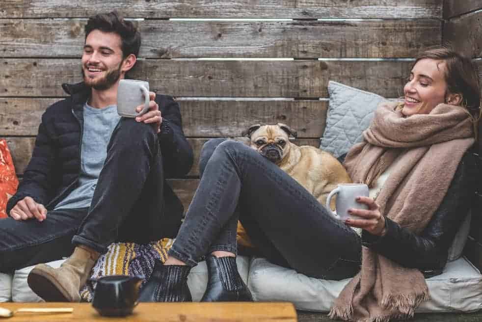Young couple hangs out with their pug while drinking coffee. To help keep your dog happy and healthy, consider feeding him CBD oil dog treats.