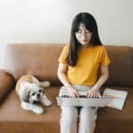 Woman sits on couch studying with her Shihi Tzu. Bringing your dog to college helps you get better grades by making you set a schedule and be more responsible.