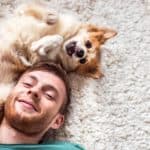 The man cuddles with the puppy while it is lying on the ground.  As a new dog parent, it is his responsibility to ensure the nutrition, health and entertainment of his dog.