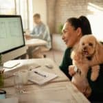 Woman holds poodle as she works in her office. Use technology, pet sitters, and dog-walking services to help create a work/life balance as a pet parent and entrepreneur.