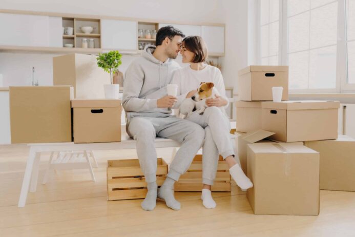 Couple sits with Jack Russell terrier while surrounded by moving boxes. 