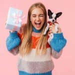 Excited woman holds gift and French bulldog puppy. Splurging on your dog can be a guilty pleasure you can indulge in. Consider these dog gift ideas to treat your best friend.