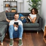 Man sits on floor using a tablet while a woman sits on the couch with two dogs while looking at her smart phone. Living with pets poses challenges for techy pet lovers. Pet parents can protect their gadgets with proper training and home set-ups.
