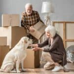 Older couple with yellow Labrador packs boxes to prepare for long-distance move.