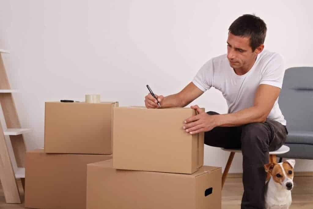 Owner labels boxes for long-distance move while his Jack Russell Terrier sits by his leg.
