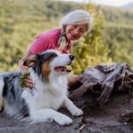 A woman pets Australian Shepherd during a rest break on a hike. The best way to prepare for dog hiking trips is to take your dog on long daily walks to ensure she is healthy and able to handle the strain of hiking.